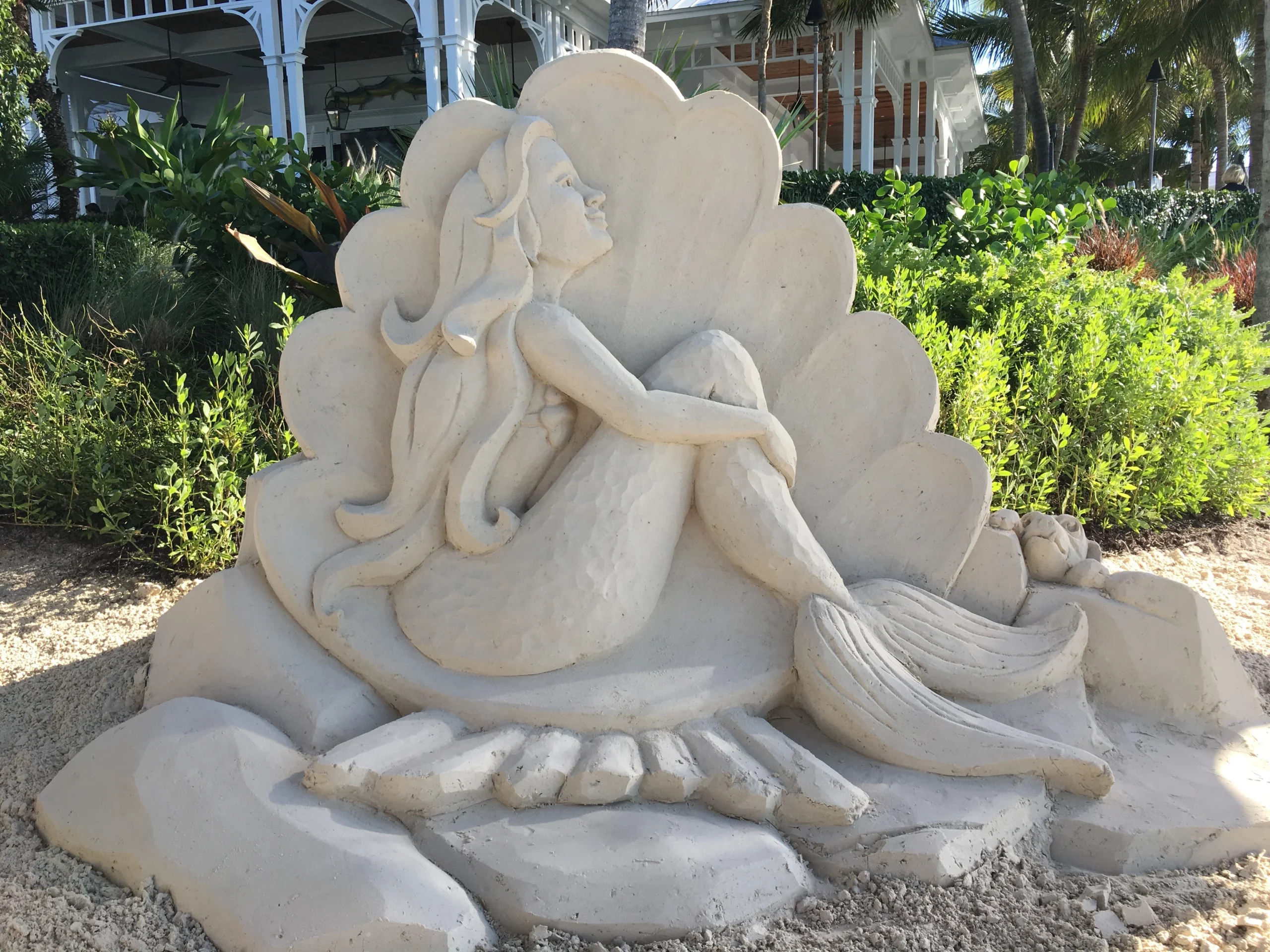 A photo of a sand sculpture of a beautiful mermaid sitting in a clamshell, looking off into the distance. It was made in front of a Florida restaurant with historic architecture.