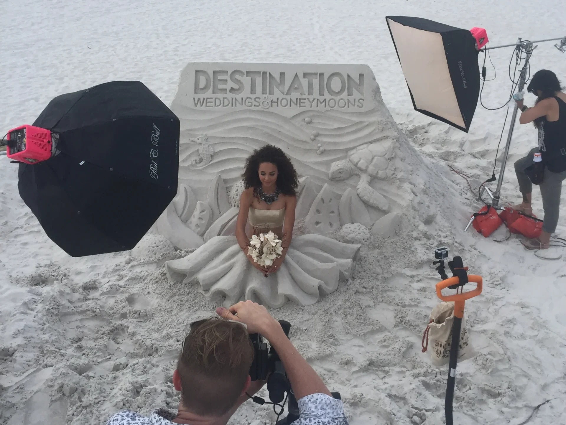 A photo of a professional photo shoot in front of a sand sculpture. The model is a woman who is sitting on the beach, her legs covered in sand, sculpted to look like a skirt. There are box lights and a photographer in the periphery of the photo. The background sand sculpture is a budget friendly flat underwater seascape with a turtle and a sea horse reading “Destination weddings and honeymoons" at the top.