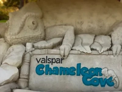 A photo of a sand sculpture of a large chameleon sitting on top of greenery and flowers above a large sign that reads “Valspar Chameleon Cove”. The letters of the sign are done in vibrant colored sand art.